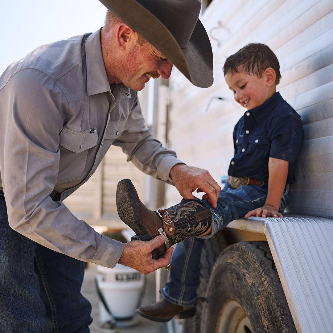 A man in a cowboy hat and western shirt helps a young boy put on a brown western boot while the boy sits on a vehicle tire against a white siding wall.