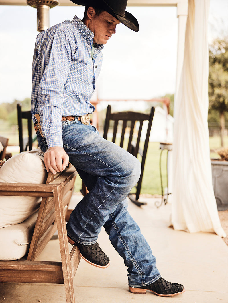 Person wearing a cowboy hat, checkered shirt, and jeans sitting on a wooden chair outdoors.