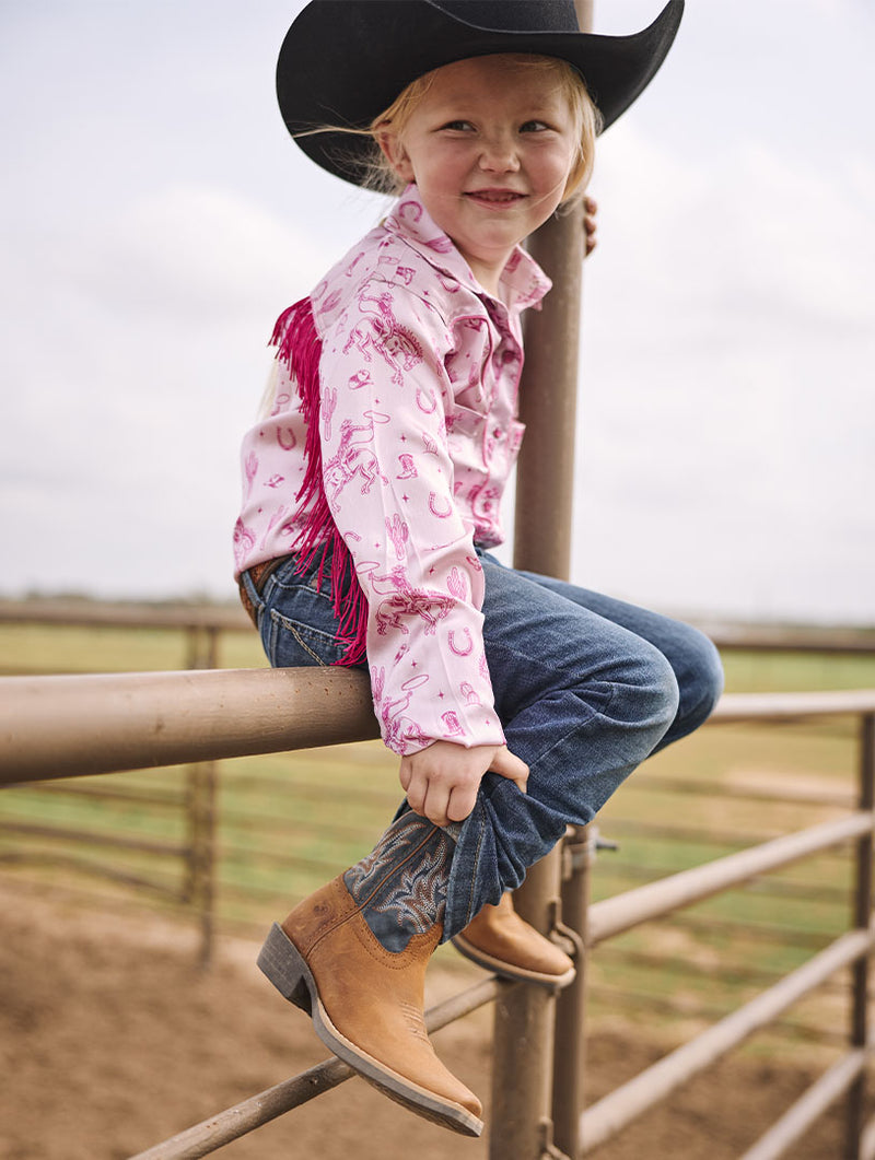 A young child wearing a pink western-style long-sleeve shirt with pink fringe, blue jeans, tan cowboy boots, and a black cowboy hat, sitting on a metal fence in an outdoor ranch setting.