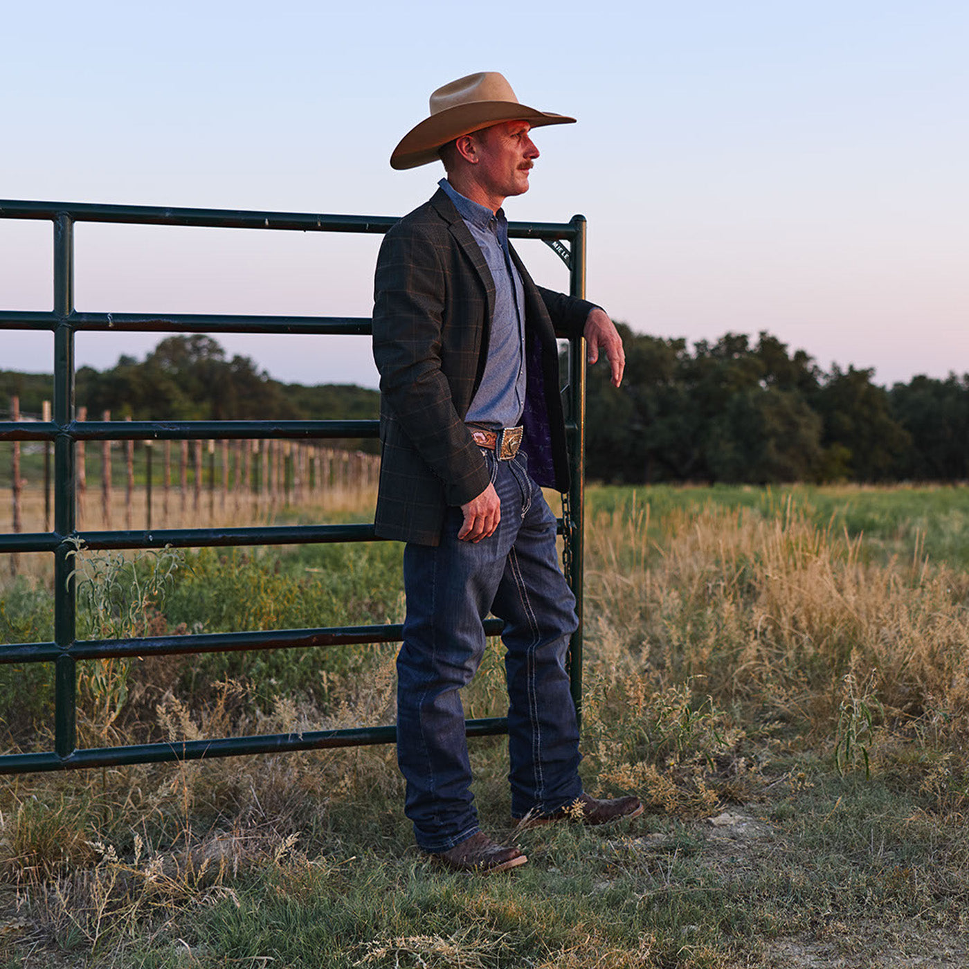 Man in cowboy hat and ostrich boots standing by a gate in a field