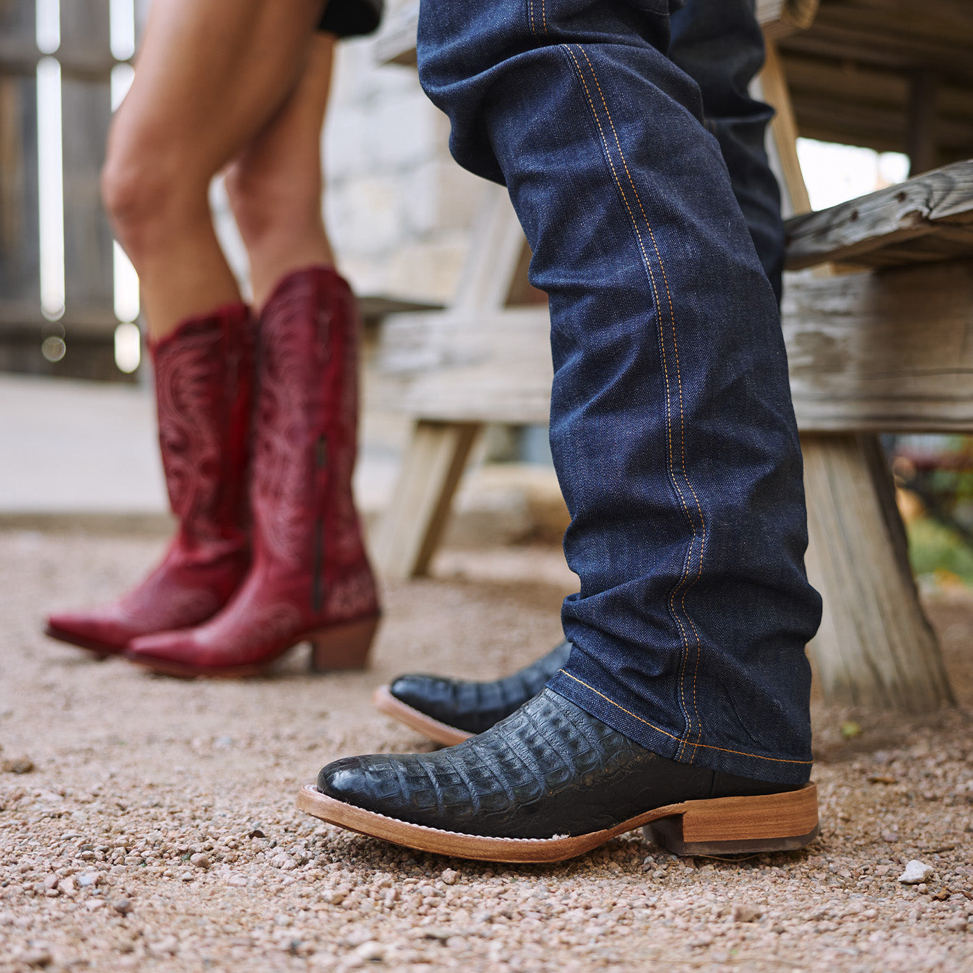 Close-up of a person wearing blue jeans and black western boots, with another person in red cowgirl boots in the background.
