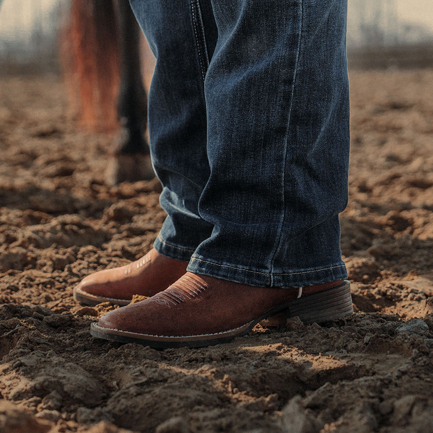 Close-up of feet wearing brown cowgirl boots and blue jeans standing on a dirt field.