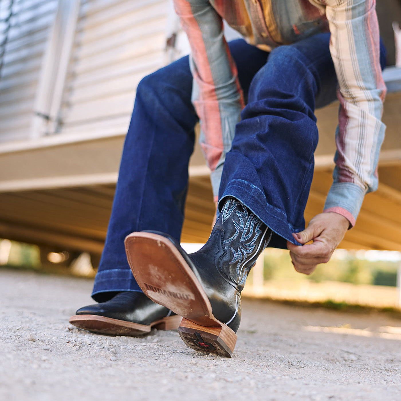 Woman wearing black cowboy boots and blue jeans with a plaid shirt, on a gravel surface.
